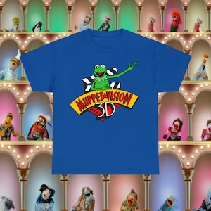 Puede incluir: Una camiseta azul real con un personaje verde de Muppet, Kermit la Rana, que lleva una estrella roja. El texto "Muppet Vision 3D" está en una pancarta amarilla con letras rojas.