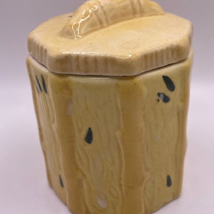 May include: A yellow ceramic jar with a lid shaped like a log. The jar has a wood grain pattern and black accents.