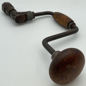 May include: A vintage hand drill with a wooden handle and a metal crank. The drill is made of metal and has a brown wooden handle. The drill is in good condition and has a patina.
