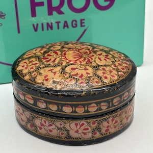 May include: A round black lidded box featuring intricate hand-painted floral patterns in shades of gold, red, and pink. The lid and the base are decorated with detailed botanical motifs, and the side of the lid displays a repeating circular pattern.