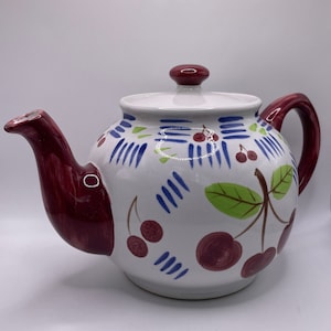 May include: A white ceramic teapot with a burgundy lid and handle. The teapot is decorated with a cherry motif in shades of red, green, blue, and white.
