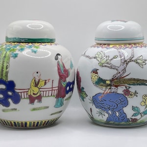 May include: Two white porcelain jars with lids, decorated with colourful floral and bird designs. One jar features a scene with a tree, a person, and a child. The other jar features a pheasant perched on a rock.