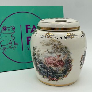 May include: A cream-coloured ceramic jar with a lid, adorned with a gold rim and floral patterns. The jar displays a detailed illustration of figures in a garden scene. A teal box with the text "FA" and a frog illustration is in the background.