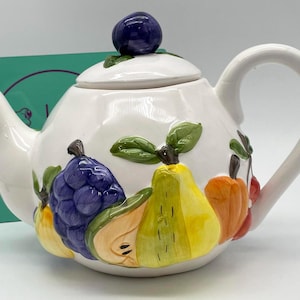 May include: White ceramic teapot with a lid topped with a purple berry. The teapot is decorated with colorful, raised relief fruit including grapes, pears, and cherries. The handle and spout are white.