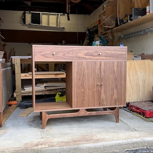 May include: A walnut wood sideboard featuring a rectangular frame with one open shelf on the left side, two drawers at the top, and two cabinet doors on the right. The piece includes four tapered legs and small, round, gold-toned metal knobs.