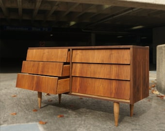 May include: A mid-century modern dresser with four drawers made of wood. The dresser is in a light brown color and has a slightly distressed finish. The drawers are open, revealing the interior of the dresser.