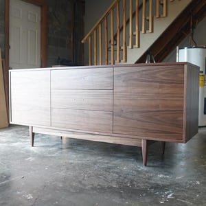 May include: A mid-century modern walnut wood sideboard with three drawers and two doors. The sideboard has tapered legs and a simple design.