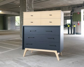 May include: A two-tone dresser with a light wood top and dark blue bottom. The dresser has six drawers with black handles. The dresser is standing on a concrete floor.
