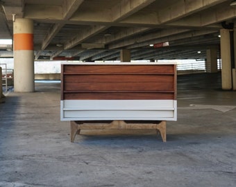 May include: A mid-century modern dresser with four drawers. The top three drawers are made of wood with a dark brown stain. The bottom drawer is white. The dresser is sitting on a concrete floor.