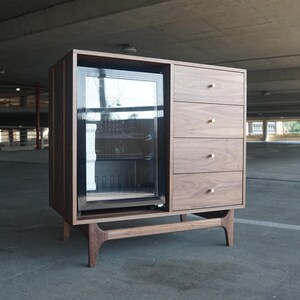 May include: A wooden bar cabinet with a built-in mini-refrigerator and four drawers. The cabinet is made of dark wood and has a modern design with gold-colored knobs on the drawers. The refrigerator has a glass door.