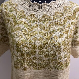 May include: A beige and green knitted sweater with short sleeves. The sweater features a detailed pattern in shades of green and beige, with a delicate, scalloped neckline. The overall design is intricate and handmade, showcasing a textured, cozy aesthetic.