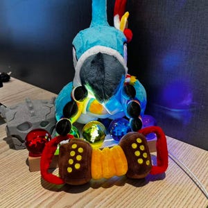 May include: A plush blue parrot with a black beak and a red and yellow crest. It has a necklace of black beads and is surrounded by colourful glass spheres and a brown and orange accordion-like toy with red handles.