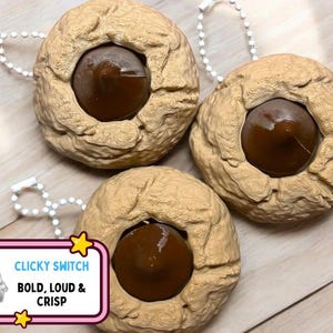 Puede incluir: Tres llaveros beige con forma de galleta con un centro marrón oscuro. Cada llavero tiene una pequeña cadena plateada adjunta. Un gráfico con el texto "CLICKY SWITCH, BOLD, LOUD & CRISP" está en la esquina inferior izquierda.