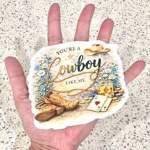 May include: A sticker with a cowboy theme. The design includes a cowboy hat, boots, a glass of whiskey, playing cards, and the text "YOU'RE A Cowboy LIKE ME" in a decorative font. The background features flowers and a rope.