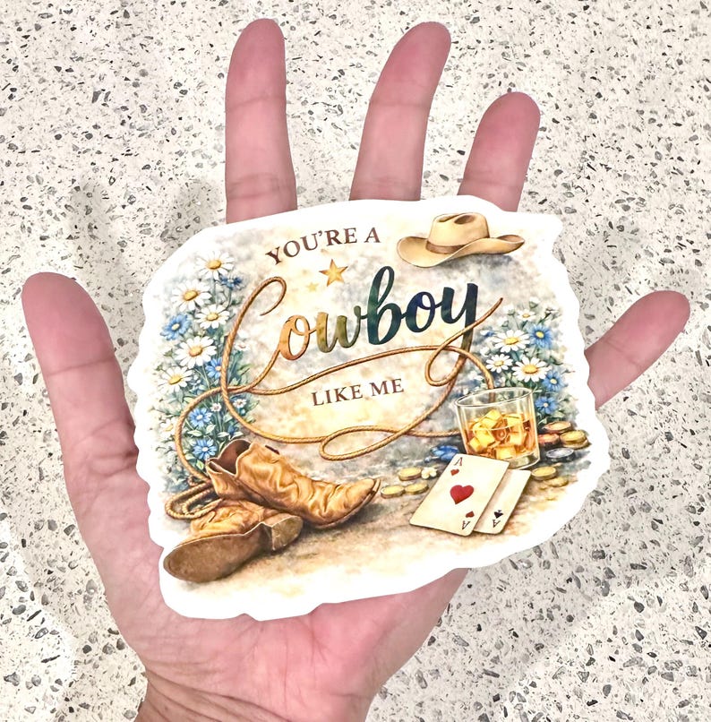 May include: A sticker with a cowboy theme. The design includes a cowboy hat, boots, a glass of whiskey, playing cards, and the text "YOU'RE A Cowboy LIKE ME" in a decorative font. The background features flowers and a rope.