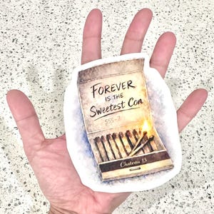 May include: A sticker featuring a matchbox design with the text "Forever is the Sweetest Con." The matchbox is open, with several matches and two lit matches. The sticker has a white border.