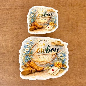 May include: Two stickers with a cowboy theme. Each sticker features a cowboy hat, boots, playing cards, and a glass of whiskey. The text "You're a Cowboy Like Me" is written in a decorative font, surrounded by flowers.