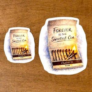 May include: Two stickers on a wooden surface. One sticker shows a matchbox with the text "FOREVER IS THE Sweetest Con" and "Chateau 13". The other sticker is a smaller version of the same image.