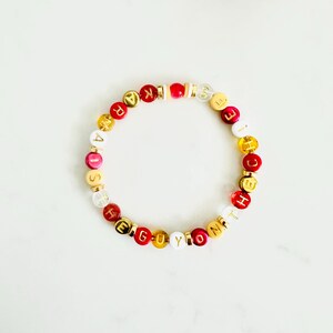 Könnte beinhalten: Ein rotes, weißes und goldenes Perlenarmband mit den Buchstaben "HE IS THE GUY YOU KNOW".