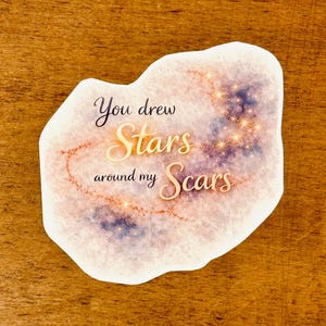 May include: A sticker with a watercolor design and the text "You drew Stars around my Scars." The sticker has a light background with a galaxy design in shades of purple and gold. The text is in a handwritten style.
