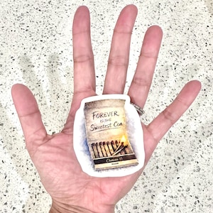 May include: A sticker featuring a matchbox design with the text "Forever is the Sweetest Con." The matchbox is tan with a partially lit match and the text "Chateau 13" at the bottom. The sticker is held in a hand.