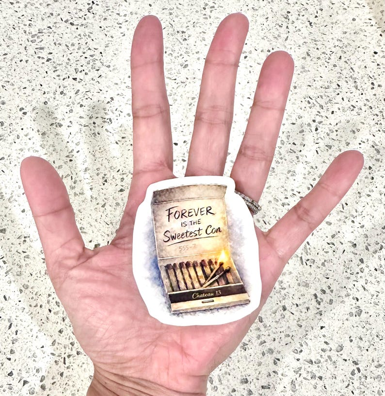 May include: A sticker featuring a matchbox design with the text "Forever is the Sweetest Con." The matchbox is tan with a partially lit match and the text "Chateau 13" at the bottom. The sticker is held in a hand.
