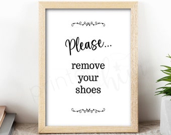 May include: A framed sign with the text "Please... remove your shoes" in black cursive font on a white background. The sign is in a light wood frame and is sitting on a table.