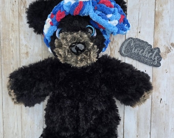 Puede incluir: Un oso de peluche negro hecho a mano con una diadema de ganchillo azul, roja y blanca. El oso tiene un hocico y patas de color marrón claro. La imagen incluye un pequeño letrero que dice "Crochets".