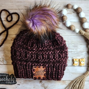 May include: A burgundy knitted beanie with a faux fur pom pom in shades of purple and brown. The beanie has a brown leather patch with the text "Sweater Weather" on it.