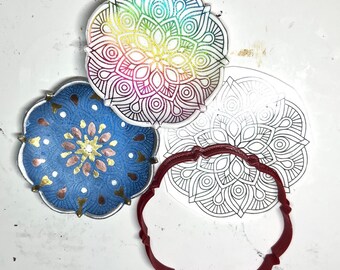Könnte beinhalten: Drei Mandala-Designs, eines mit Regenbogenfarben, eines mit goldenen und kupfernen Akzenten auf blauem Hintergrund und eines mit einer leeren Mandala-Umrandung. Die Designs befinden sich auf einer weißen Oberfläche.