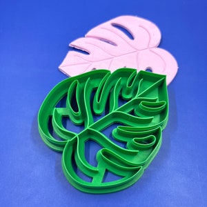 Könnte beinhalten: Zwei Ausstecher in Form eines Monstera-Blattes. Der obere Ausstecher ist rosa und der untere ist grün.