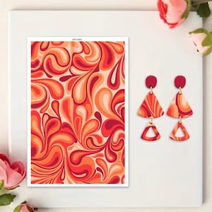 Pode incluir: Em um fundo branco, uma impressão emoldurada apresenta um padrão abstrato e rodopiante em tons de vermelho, laranja e pêssego. Dois pares de brincos com um design triangular e detalhes em vermelho também são visíveis.