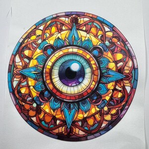 Puede incluir: Un mandala de vitral colorido con un gran ojo azul en el centro. El ojo tiene un iris morado y una pupila blanca. El mandala está rodeado por un patrón intrincado de rojo, naranja, amarillo, azul y verde.