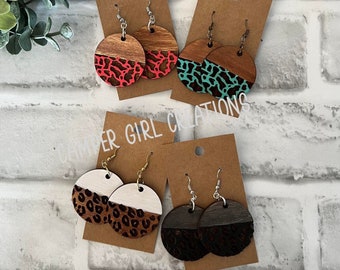 May include: Four pairs of wooden earrings with a half-circle design. The earrings are painted with different animal print patterns, including leopard print, pink cheetah print, and turquoise cheetah print. The earrings are on brown card stock with the text "CAMPER GIRL CREATIONS" printed on the card.