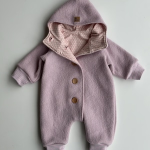 Könnte beinhalten: Helllila Baby-Overall mit Kapuze und Holzknöpfen. Die Innenseite der Kapuze und des Kragens weist ein rosa Wabenmuster auf. Die Manschetten und Knöchel haben gestreifte Details. Ein kleines Etikett auf der Kapuze lautet "OH HELLO".