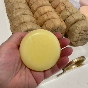 May include: A close-up of a yellow solid shampoo bar in a hand. The bar is surrounded by other wrapped shampoo bars in a white tray with a gold accent.