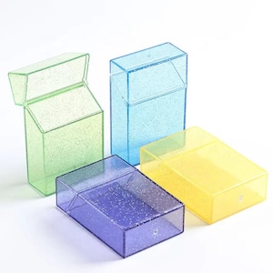 May include: Four colorful, transparent cigarette cases in green, blue, purple, and yellow. Each case has a rectangular shape with a hinged lid. The cases are slightly glittery, and the lids are closed or open.