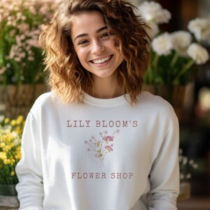 May include: A woman wearing a white sweatshirt with the text "Lily Bloom's Flower Shop" and a floral design.
