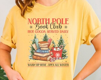 Könnte beinhalten: Ein gelbes T-Shirt mit einem Weihnachtsmotiv. Das Design zeigt einen Stapel Bücher, eine Tasse heiße Schokolade und Zuckerstangen. Der Text auf dem Shirt lautet "North Pole Book Club, Hot Cocoa Served Daily, Warm Up Here. Open All Winter."
