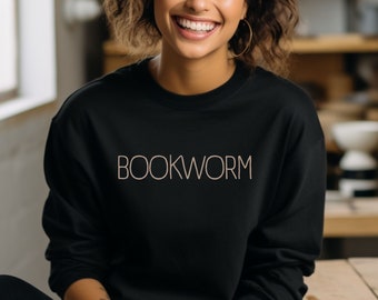 Pode incluir: Uma mulher vestindo uma camiseta preta com a palavra "BOOKWORM" impressa em marrom claro na frente. Ela está sorrindo e sentada em uma superfície de madeira.