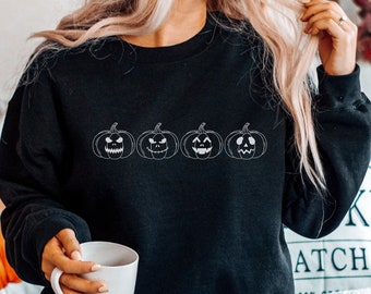 May include: Black sweatshirt with a white outline of four jack-o'-lantern faces. The sweatshirt is being worn by a person with long blonde hair.
