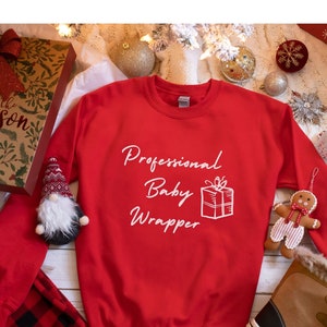 Puede incluir: Sudadera roja de Navidad con texto blanco que dice "Professional Baby Wrapper" y un dibujo de una caja de regalo.