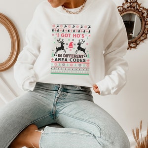 May include: White Christmas jumper with a festive design featuring reindeer and the text "I GOT HO'S IN DIFFERENT AREA CODES".