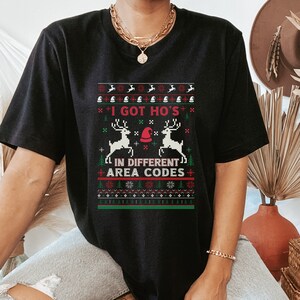 May include: Black t-shirt with a Christmas-themed graphic. The design features the text "I GOT HO'S IN DIFFERENT AREA CODES" in a pixelated, festive style with reindeer, Santa hats, and snowflakes. The shirt is a casual wear item.