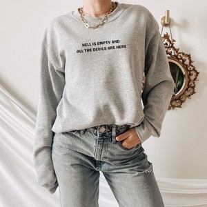 May include: A light gray sweatshirt with the text "HELL IS EMPTY AND ALL THE DEVILS ARE HERE" in black. The sweatshirt is crew neck and long-sleeved. The person is wearing light wash denim jeans.