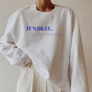 May include: White sweatshirt with the blue text "IT'S OKAY. I get lost sometimes." The sweatshirt has long sleeves and a crew neck. The model is wearing light-colored pants.