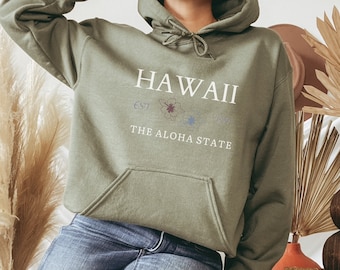 Puede incluir: Una sudadera con capucha verde con el texto "HAWAII EST 1959 THE ALOHA STATE" y un diseño floral.