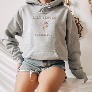 Pode incluir: Um moletom cinza claro com bolso frontal e cordão. O moletom apresenta o texto "LILY BLOOMS FLOWER SHOP" em marrom, juntamente com um gráfico floral. A modelo está usando shorts jeans.
