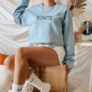 Puede incluir: Una sudadera de color azul claro con la palabra "IGNITE" impresa en negro en la parte delantera. La sudadera la lleva puesta una persona sentada en una caja de madera. La persona lleva calcetines blancos con logotipos de Nike negros.