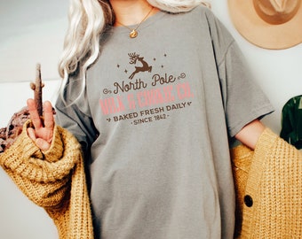 May include: A gray t-shirt with a brown and pink graphic design that says "North Pole Milk & Cookie Co." and "Baked Fresh Daily - Since 1842 -"